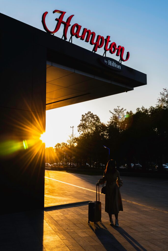 A traveler with luggage arrives at a Hampton by Hilton hotel during sunset, highlighting the silhouette against the warm sky.
